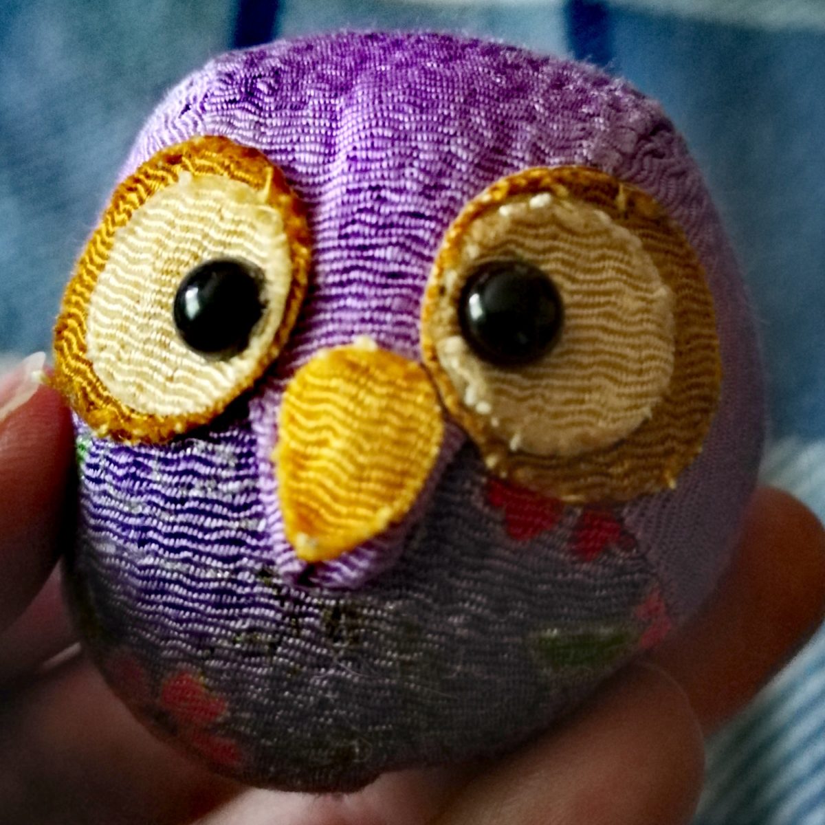 A little purple owl toy that is mostly round and sits nicely in the palm of your hand. Its bulk is made in a plain lilac purple, with a section of fabric formed into a triangular point that makes the owls face and feet. Little Owls belly is marked by another piece of fabric that has a slightly darker background, and is covered in silver swirls and pink and white and red cherry blossoms. The owl's face is finished with fabric that has been glued on rather than sewn on. Its eyes are made from concentric circles of brown, with small, lighter pieces off centre for the larger circles. The smaller dark pieces give the eyes a new moon sort of look. The pupils are black matte plastic and look straight ahead. The beak is marked by a piece of orange yellow fabric. It is held in the left hand of a white woman. The background is a white and blue plaid fleece blanket.